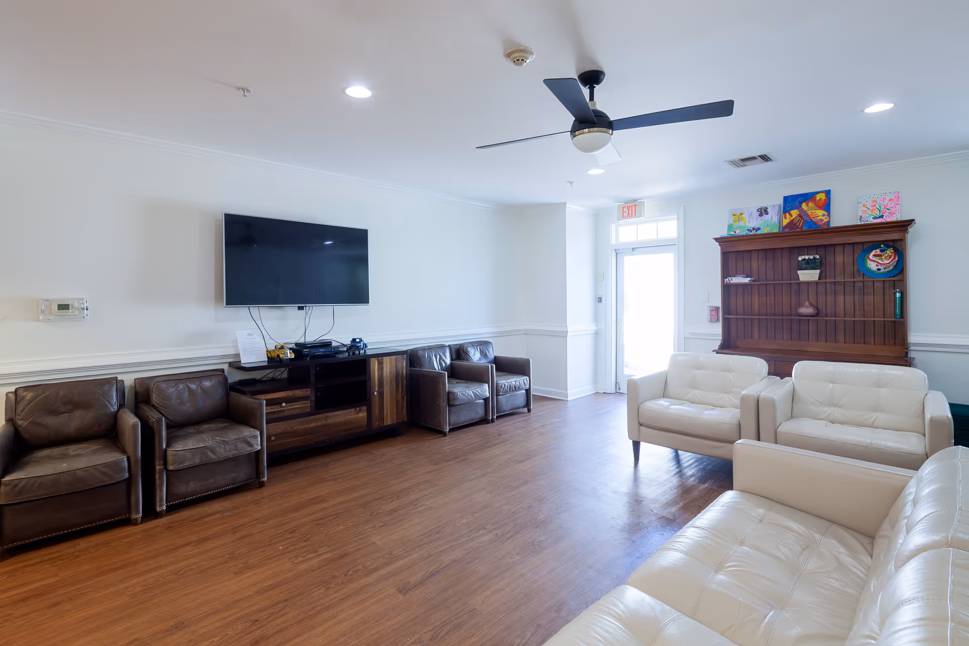 A bright living room with wooden flooring, featuring a wall-mounted flat screen TV above a wooden media console. There are four dark brown leather armchairs lined up along the left wall and three white leather sofas arranged on the right side. A ceiling fan with lights is mounted on the white ceiling. A wooden hutch with colorful artwork and decorative items is against the far wall near a door with an exit sign above it.