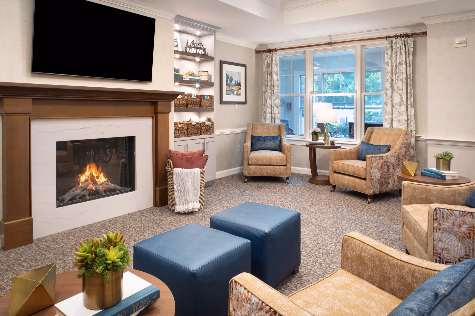 A cozy living room with a lit fireplace, a mounted flat-screen TV above it, and comfortable armchairs arranged around two blue ottomans. There are side tables with decorative items and a large window with patterned curtains letting in natural light.