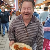 An older adult smiling and holding a plate of food in a cafeteria-style dining area with other people in the background.