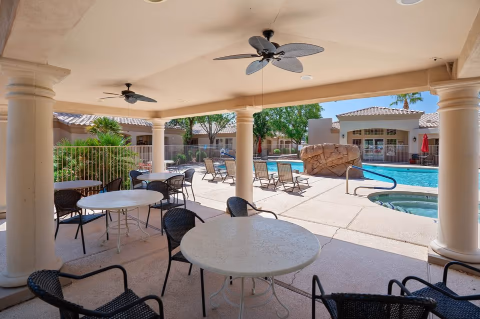 Covered outdoor seating area with round tables and black chairs overlooking a swimming pool with lounge chairs, a large rock feature, and a hot tub. The area is surrounded by a fence and buildings with tiled roofs, under a clear blue sky.