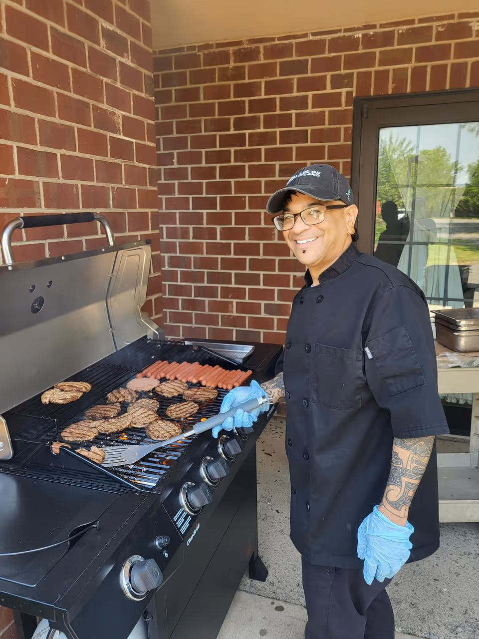 A man wearing a black chef coat, black cap, glasses, and blue gloves is grilling hamburgers and hot dogs on a large outdoor gas grill. He is smiling and holding a spatula. The background shows a brick wall and a glass door reflecting outdoor greenery.