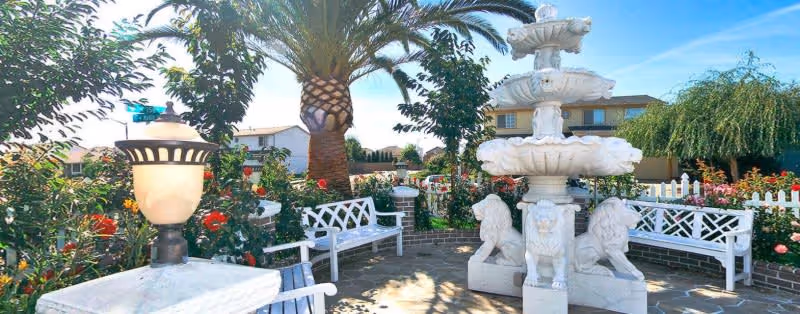 Sunny outdoor courtyard with a white multi-tiered fountain flanked by lion sculptures, white benches, a palm tree and flowering shrubs.