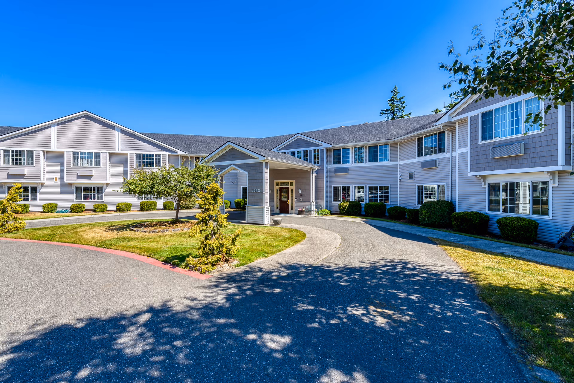 Exterior view of Laurel Place Assisted Living & Memory Care building on a sunny day, showing a two-story structure with multiple windows, a covered entrance, and a driveway surrounded by green lawns and small trees.