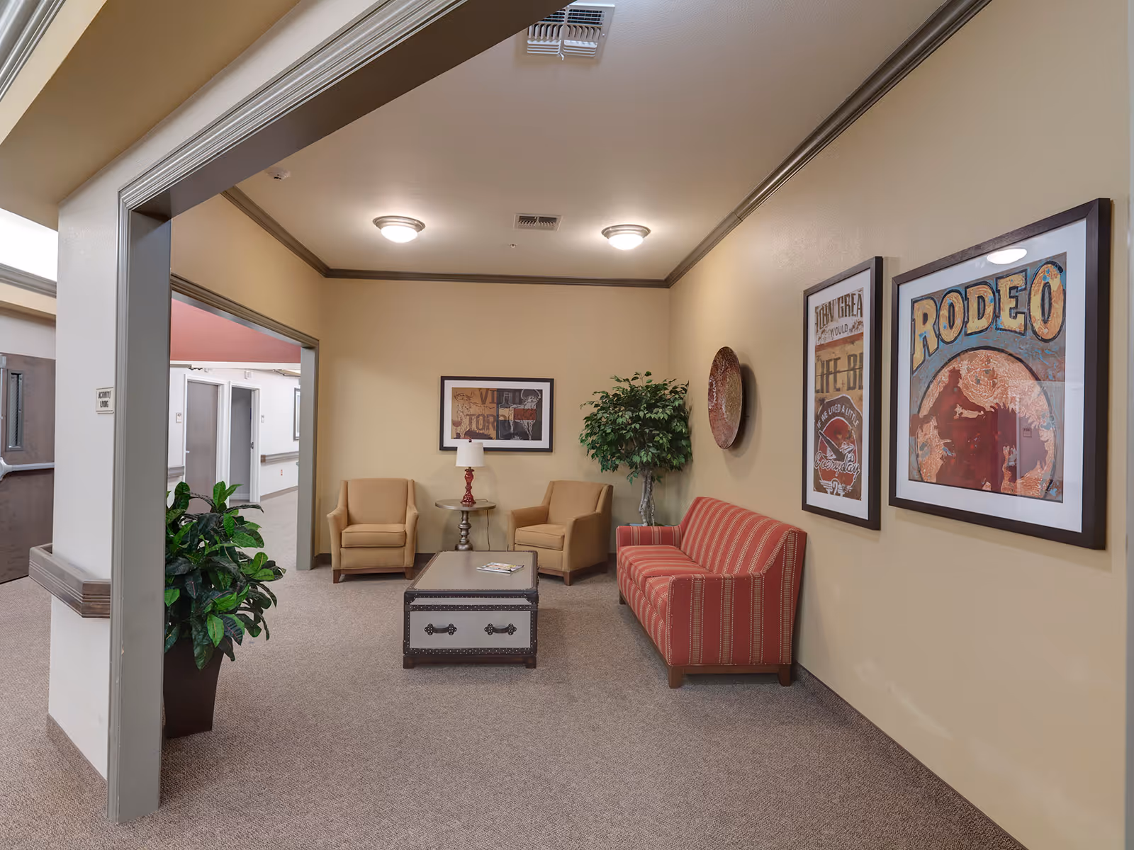 A cozy sitting area with two armchairs, a striped sofa, a coffee table, framed wall art, and potted plants in a senior living facility corridor.