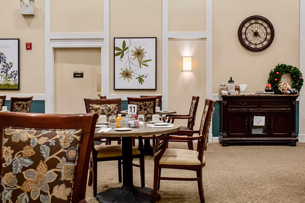 Dining area with round tables and wooden chairs featuring floral upholstery. Tables are set with plates, cups, glasses, and condiments. On the wall, there are framed botanical prints, a large decorative clock, and a sideboard with jars, napkins, and a Christmas wreath. A door labeled 'Kitchen' is visible in the background.