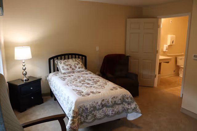 A single bed with a floral quilt and matching pillow in a bedroom. Next to the bed is a black nightstand with a lamp. There is a cushioned armchair with a dark red throw blanket draped over it. An open door reveals a bathroom with a toilet, sink, and paper towel dispenser. The room has beige walls and carpeted floor.