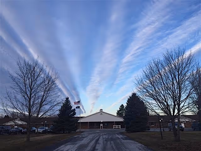 Exterior view of Medilodge of Sterling building with a driveway leading to the entrance, flanked by leafless trees and evergreen trees under a blue sky with streaked clouds.