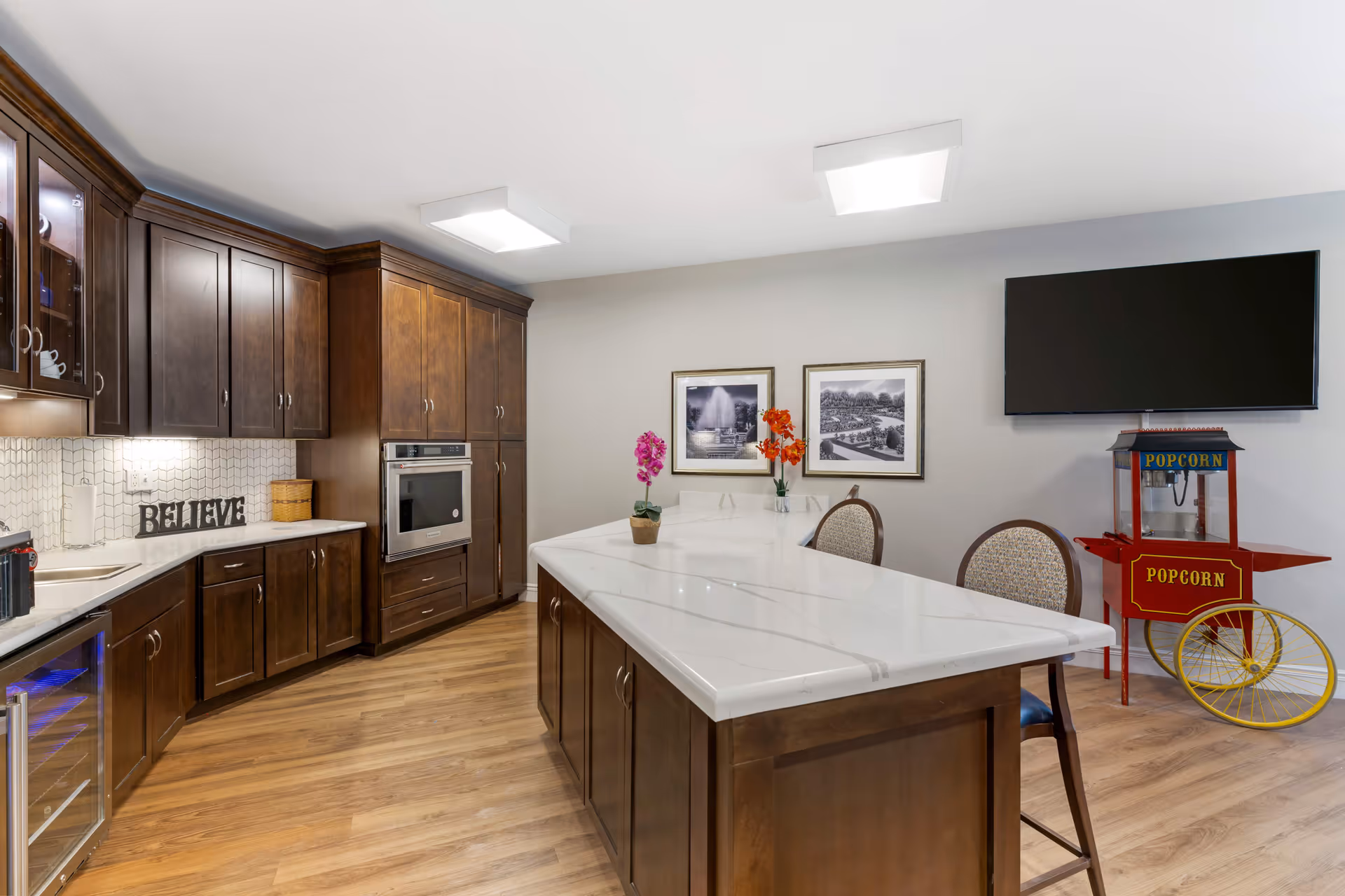 A modern kitchen area with dark wooden cabinets, a white marble countertop island with two chairs, a built-in oven, a small wine cooler, and a popcorn machine under a wall-mounted flat screen TV. The walls are light gray with two framed black and white pictures and two vases with colorful flowers on the island.