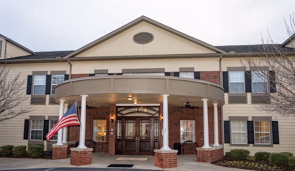 Front exterior view of The Carriages of Rivergate facility showing a two-story building with beige and brick facade, a covered entrance supported by white columns, an American flag on the left side, and some landscaping with bushes and small trees.