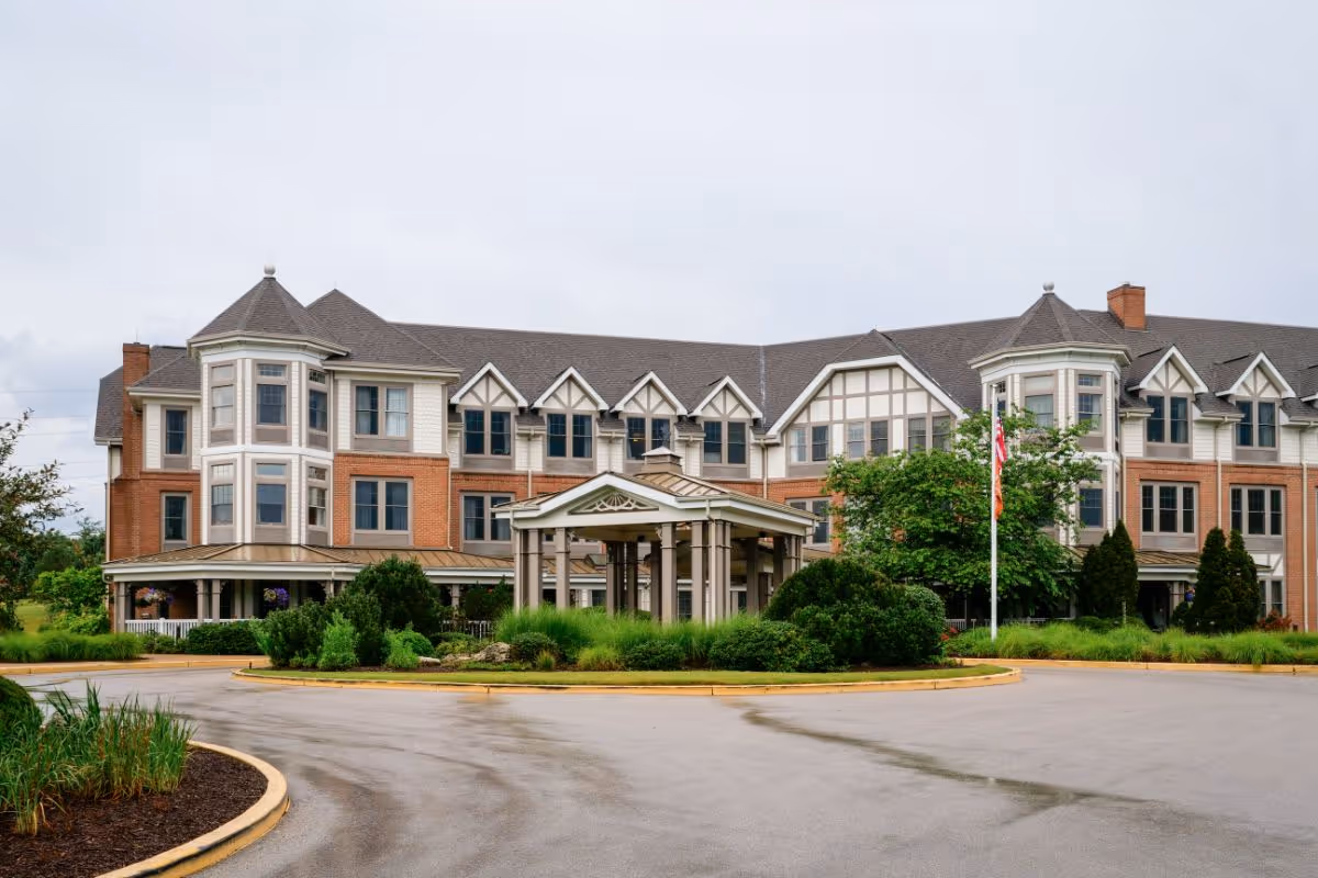 Exterior view of a large, multi-story senior living facility building with a covered entrance, surrounded by greenery and a circular driveway.