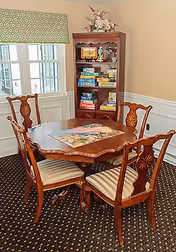 A small dining area with a wooden table and four matching chairs with striped cushions. On the table is a partially completed jigsaw puzzle. Behind the table is a wooden bookshelf filled with board games and decorative items. The room has beige walls, white wainscoting, a patterned carpet, and a window with a green patterned valance.