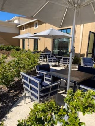 Outdoor patio area with several tables and chairs under large white umbrellas next to a beige building. Green shrubs line the walkway beside the seating area.