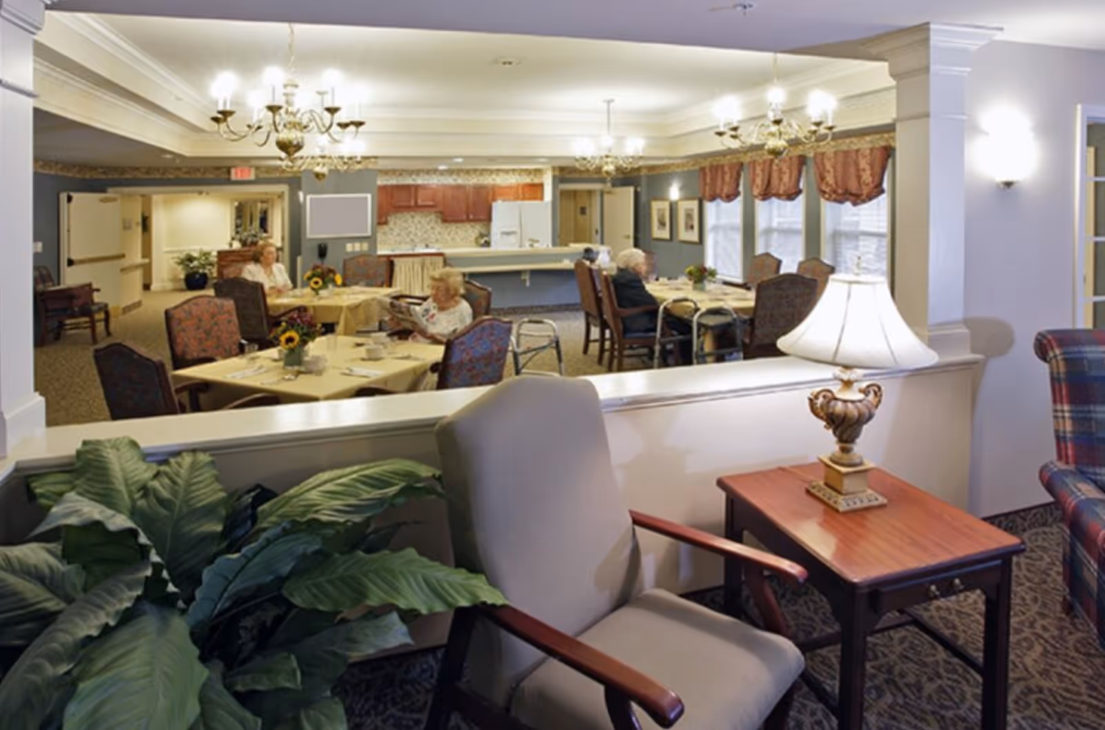 A cozy dining area in a senior living facility with several elderly residents seated at tables covered with yellow tablecloths. The room features chandeliers, a kitchen area with wooden cabinets and a refrigerator in the background, and large windows with decorative curtains. In the foreground, there is a comfortable armchair next to a wooden side table with a decorative lamp and a large green plant nearby.