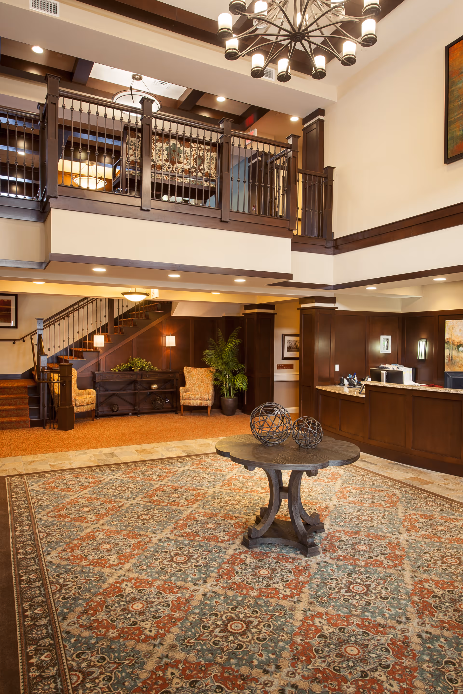 Spacious senior living facility lobby with a large patterned rug, a round wooden table with decorative metal spheres, a reception desk, two armchairs near a staircase, and a second-floor balcony with wooden railings. Warm lighting and wood paneling create a welcoming atmosphere.