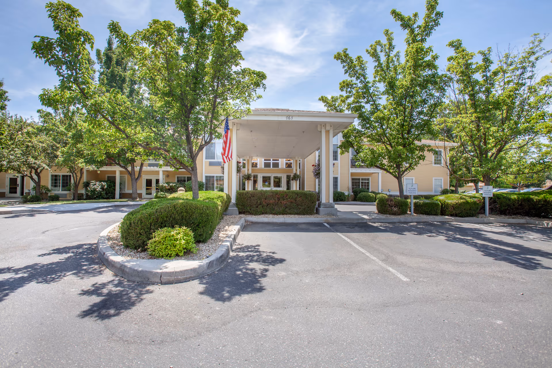 Front entrance of a two-story beige senior living building with a covered porte-cochere, American flag, trees, and parking area.