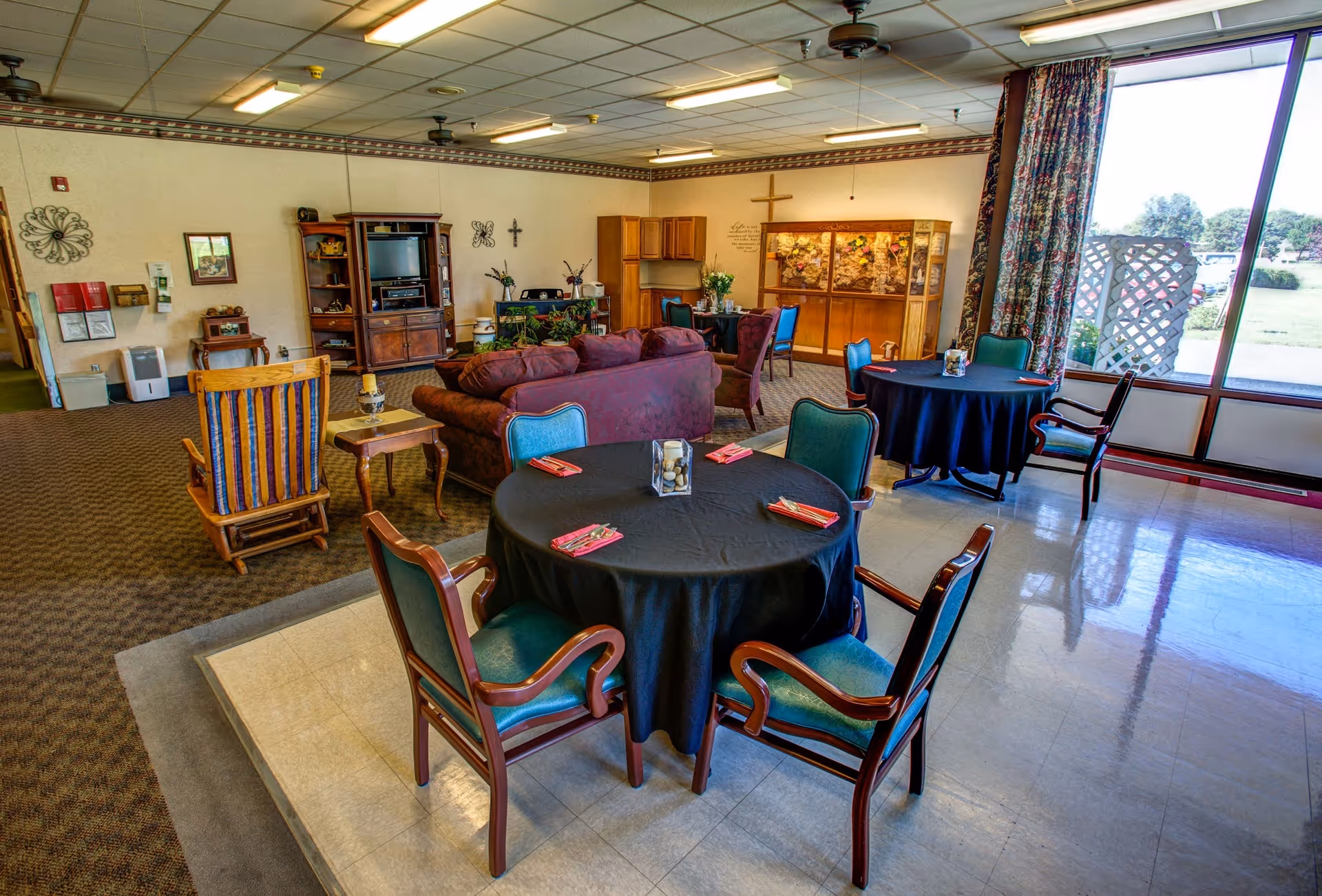 Open common room with round dining tables and chairs, a seating area with sofas and a TV, and large windows looking outside.