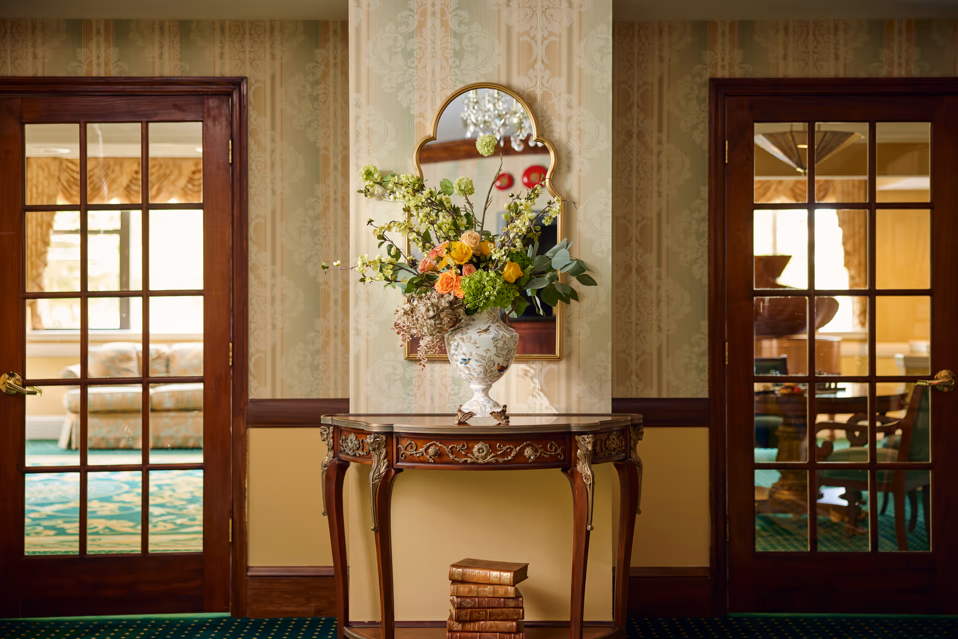 An elegant interior hallway featuring a decorative wooden table with intricate carvings, topped with a floral arrangement in a vase. Above the table is a uniquely shaped mirror. The hallway has patterned wallpaper and green carpet with a yellow and blue design. On either side of the table are wooden framed glass doors leading to other rooms with visible furniture.