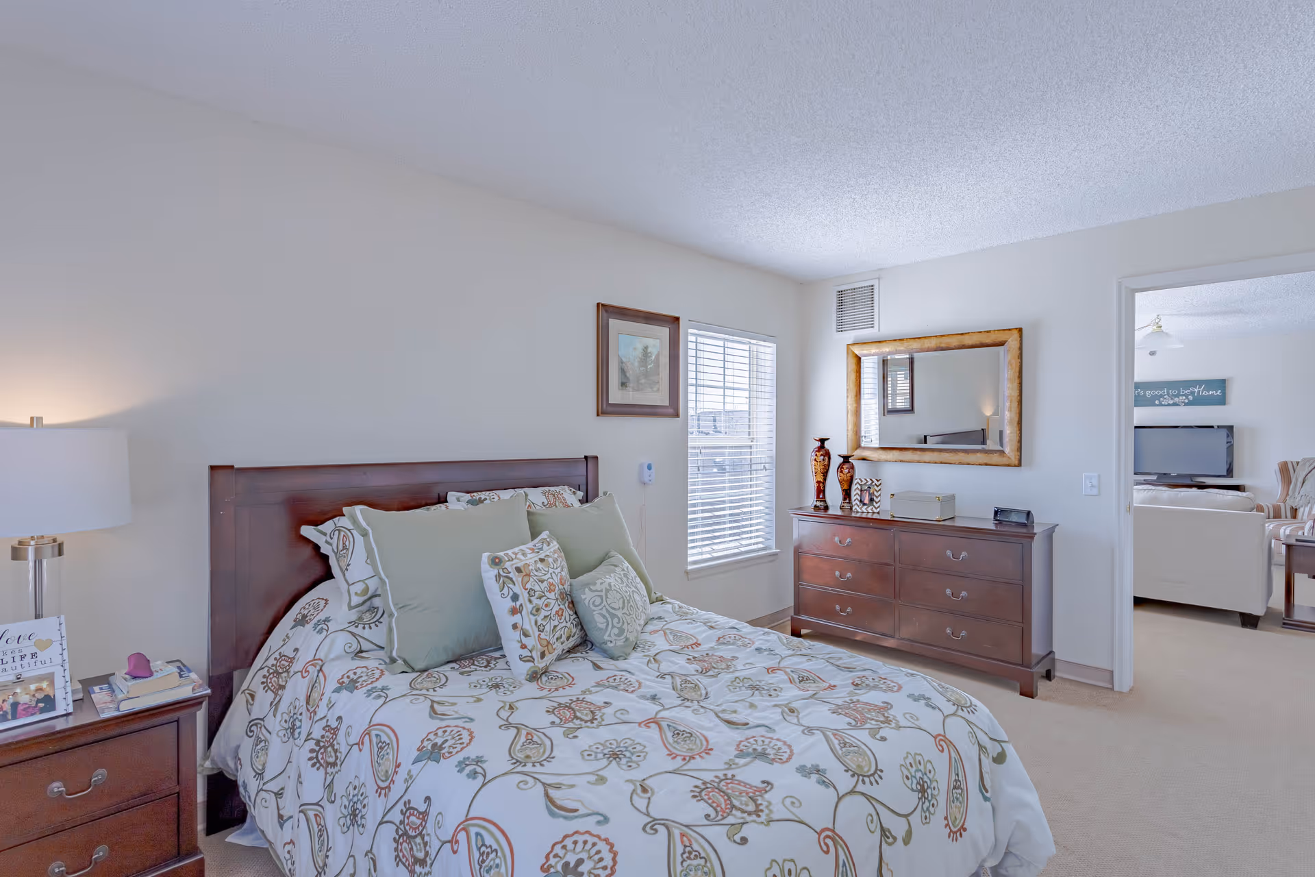 A bright and cozy bedroom with a wooden bed frame and a floral patterned bedspread with multiple pillows. There is a wooden nightstand with a lamp, books, and a framed photo. A window with blinds lets in natural light. Across from the bed is a wooden dresser with decorative vases and a large framed mirror above it. An open doorway leads to a living room area with a TV and seating visible.