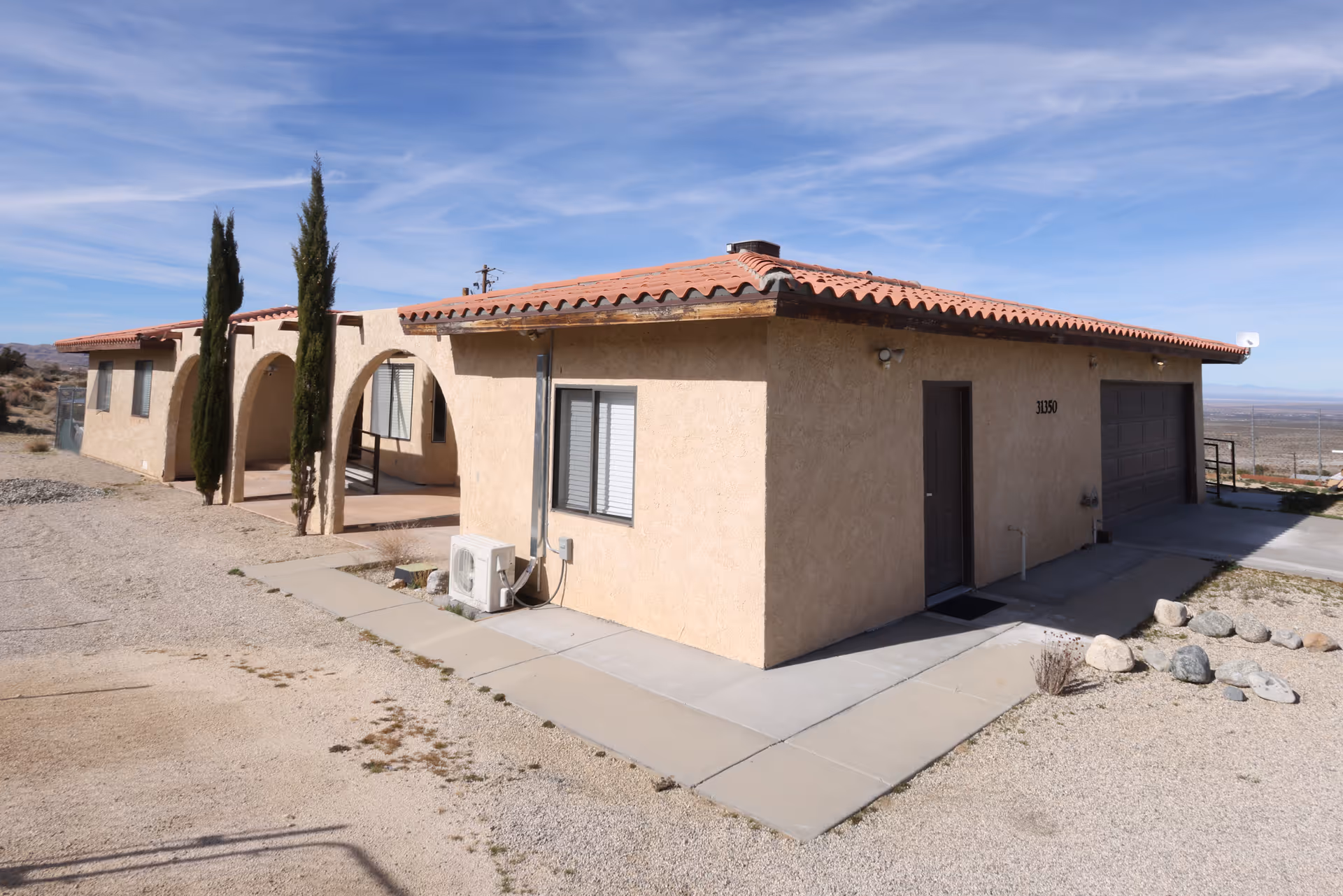 Exterior view of a single-story building with beige stucco walls and a red tile roof under a blue sky. The building has a garage door, a side door, and windows with blinds. There are two tall, narrow trees near an arched entrance, and the surrounding area is covered with gravel and rocks.