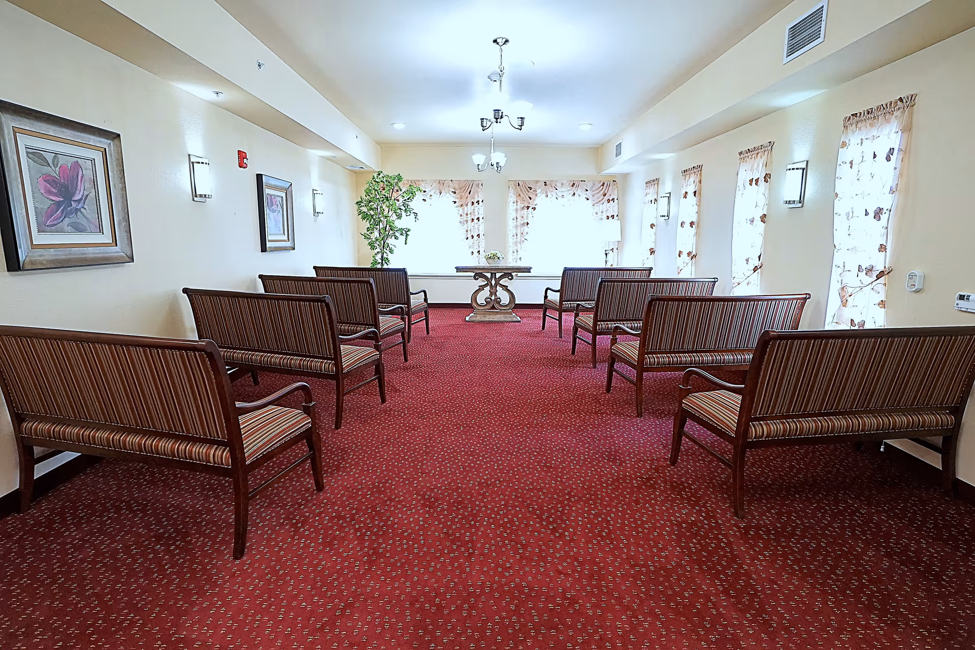 A well-lit room with multiple wooden benches upholstered in striped fabric arranged in two rows facing a small table with a decorative base at the far end. The room has a red patterned carpet, cream-colored walls adorned with framed floral artwork, several windows with sheer curtains featuring a butterfly pattern, and ceiling light fixtures.