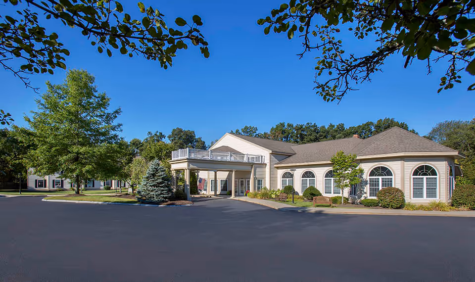Exterior view of Benchmark Senior Living at Shrewsbury Crossings showing a single-story building with large arched windows, a covered entrance, and surrounding greenery including trees and shrubs under a clear blue sky.