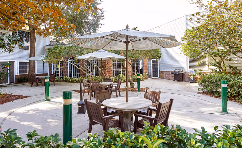 Outdoor courtyard area with round tables and chairs under large striped umbrellas surrounded by greenery and trees, adjacent to a building with white siding and brick walls.