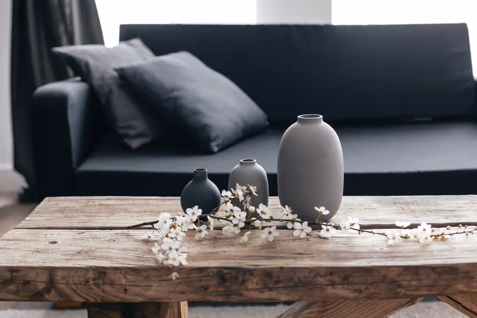 A rustic wooden coffee table with three modern vases in varying sizes and colors, accompanied by a branch of white flowers, set in front of a dark gray sofa with matching pillows in a softly lit living room.