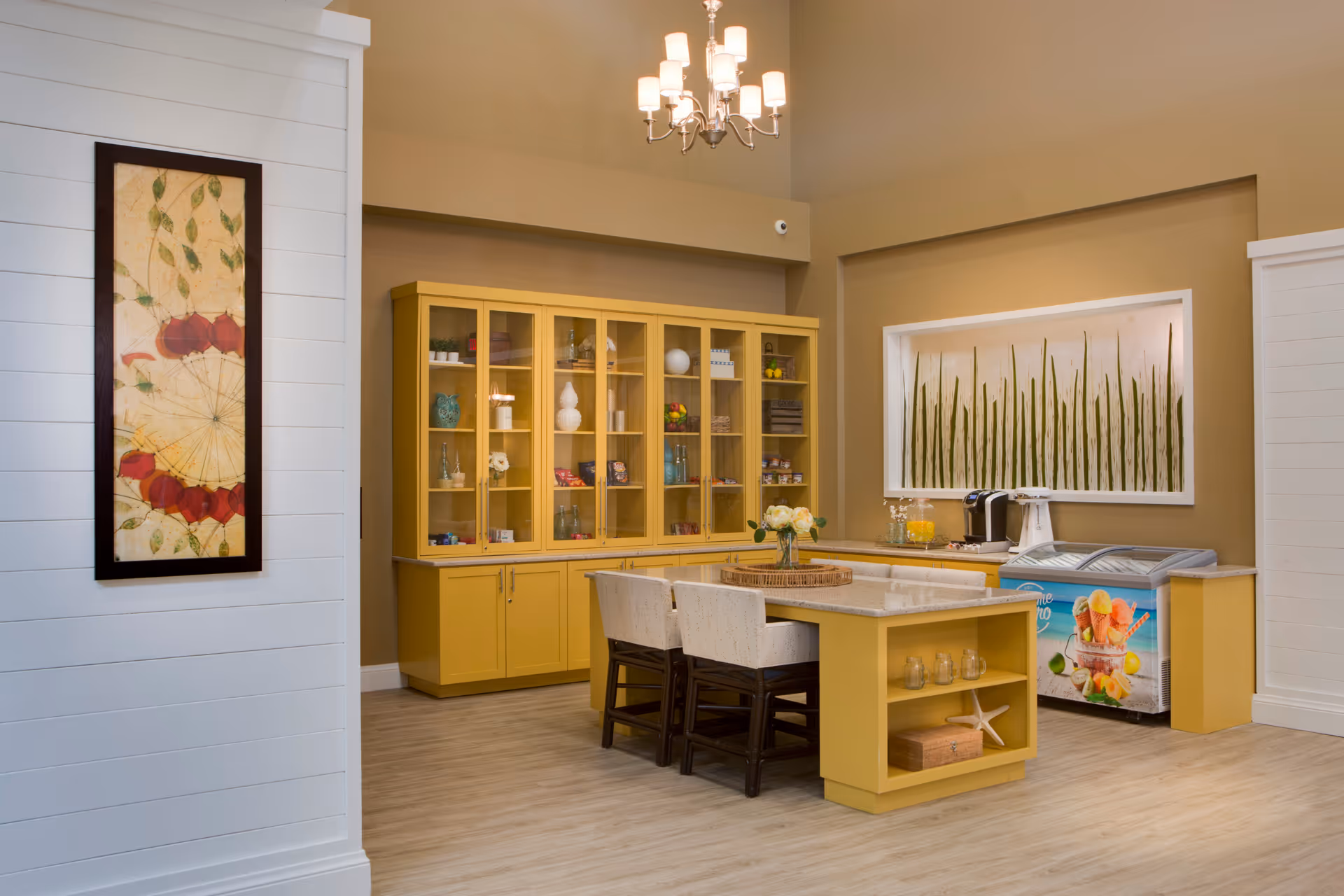 A bright and inviting interior space featuring a yellow kitchen island with two upholstered chairs, a matching yellow cabinet with glass doors displaying decorative items, a chandelier overhead, and a colorful freezer with fruit images. The walls are painted beige with white trim, and there is framed artwork on the walls.