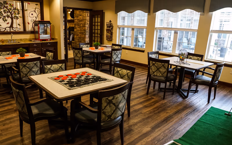 Activity/dining room with several square tables and chairs, game boards and a bingo cage set up near large windows.