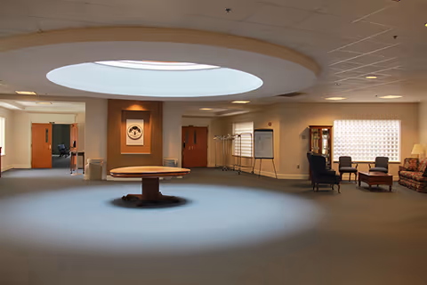 A spacious interior common area with a large circular skylight in the ceiling illuminating a round wooden table in the center. The room has carpeted floors, neutral-colored walls, and several seating areas with chairs and a sofa. There is a coat rack, a whiteboard, and framed artwork on the walls.