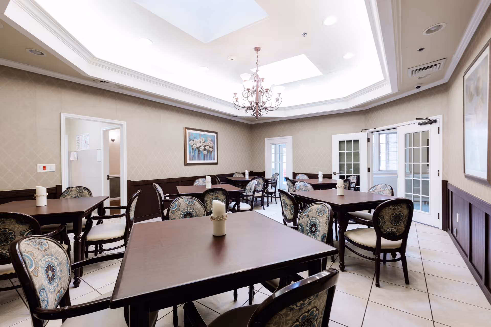 A bright dining room with several dark wooden tables and patterned cushioned chairs. The room has beige walls with a framed floral painting, a chandelier hanging from a recessed ceiling with skylights, and white tiled floors. French doors and an open doorway are visible in the background.