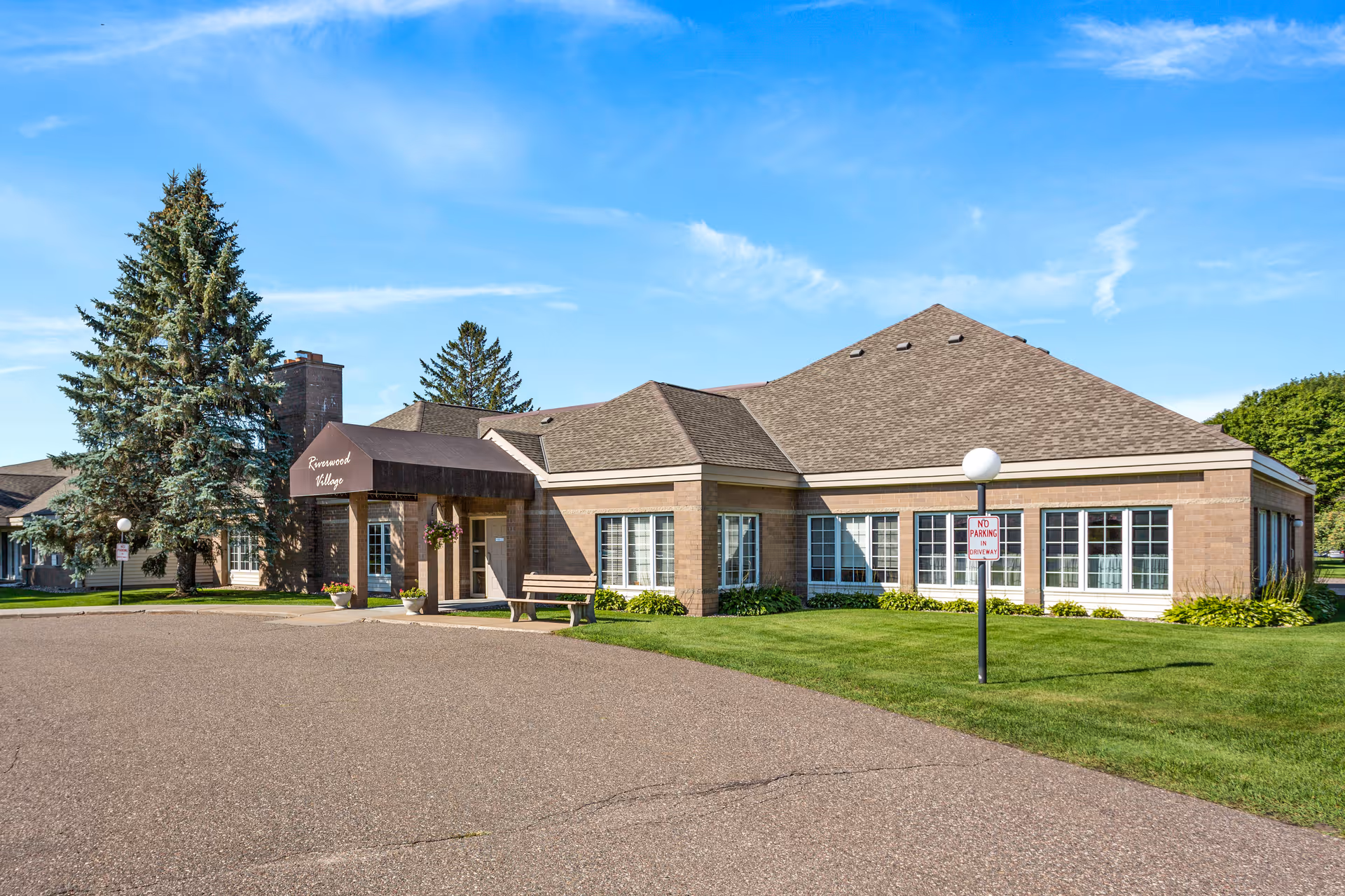 Exterior view of Riverwood Village, a single-story brick building with a brown shingled roof and a covered entrance. There are large windows along the front and side, a bench near the entrance, and a well-maintained lawn with trees and shrubs. The sky is clear and blue.