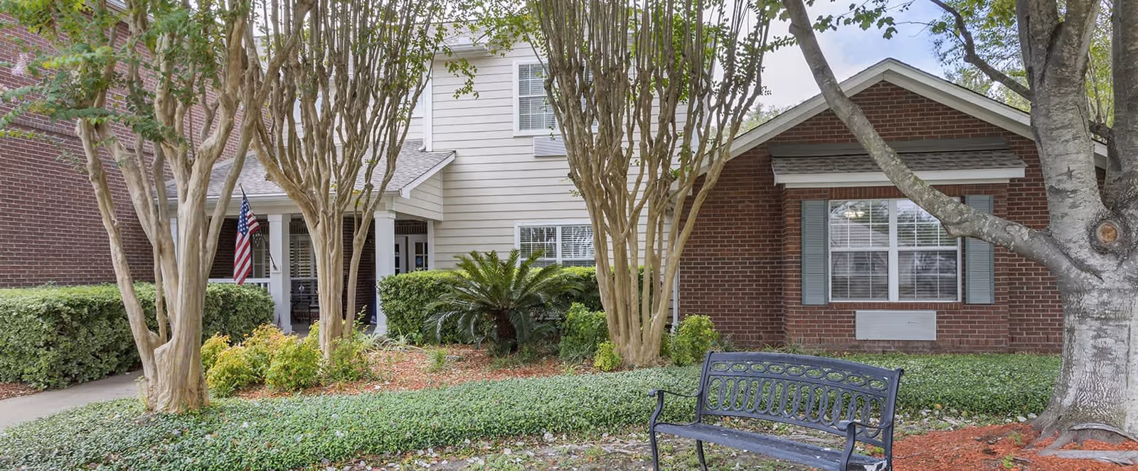 Exterior view of Brookdale Spring Shadows facility showing a brick and siding building with windows, an American flag near the entrance, landscaped bushes, trees, and a black metal bench in the foreground.