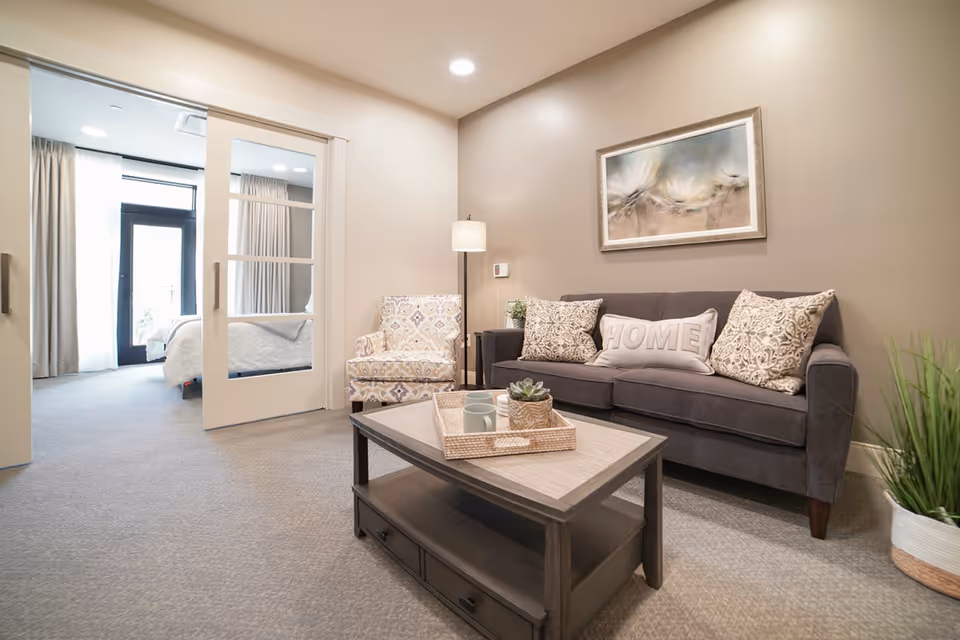 A cozy living room area with a dark gray sofa adorned with patterned pillows and a 'HOME' pillow, a patterned armchair, a wooden coffee table with a tray holding mugs and a small plant, a floor lamp, and a framed artwork on the wall. Sliding glass doors lead to a bedroom with a bed and curtains, and a door to the outside is visible in the background.
