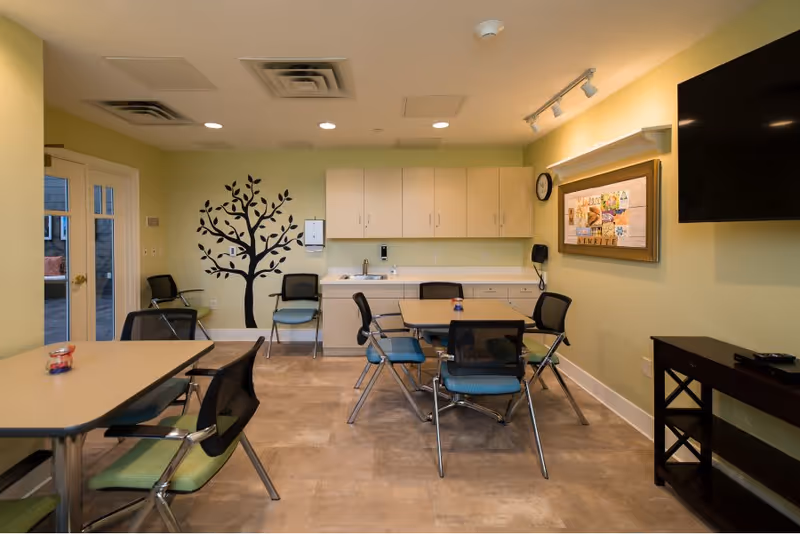 A well-lit room with light green walls featuring a black tree decal. The room contains two tables surrounded by chairs with black backs and green or blue seats. There are white cabinets and a sink along the back wall. A clock and a framed bulletin board are mounted on the right wall, and a large flat-screen TV is mounted above a dark wooden console table. Double glass doors are visible on the left side.