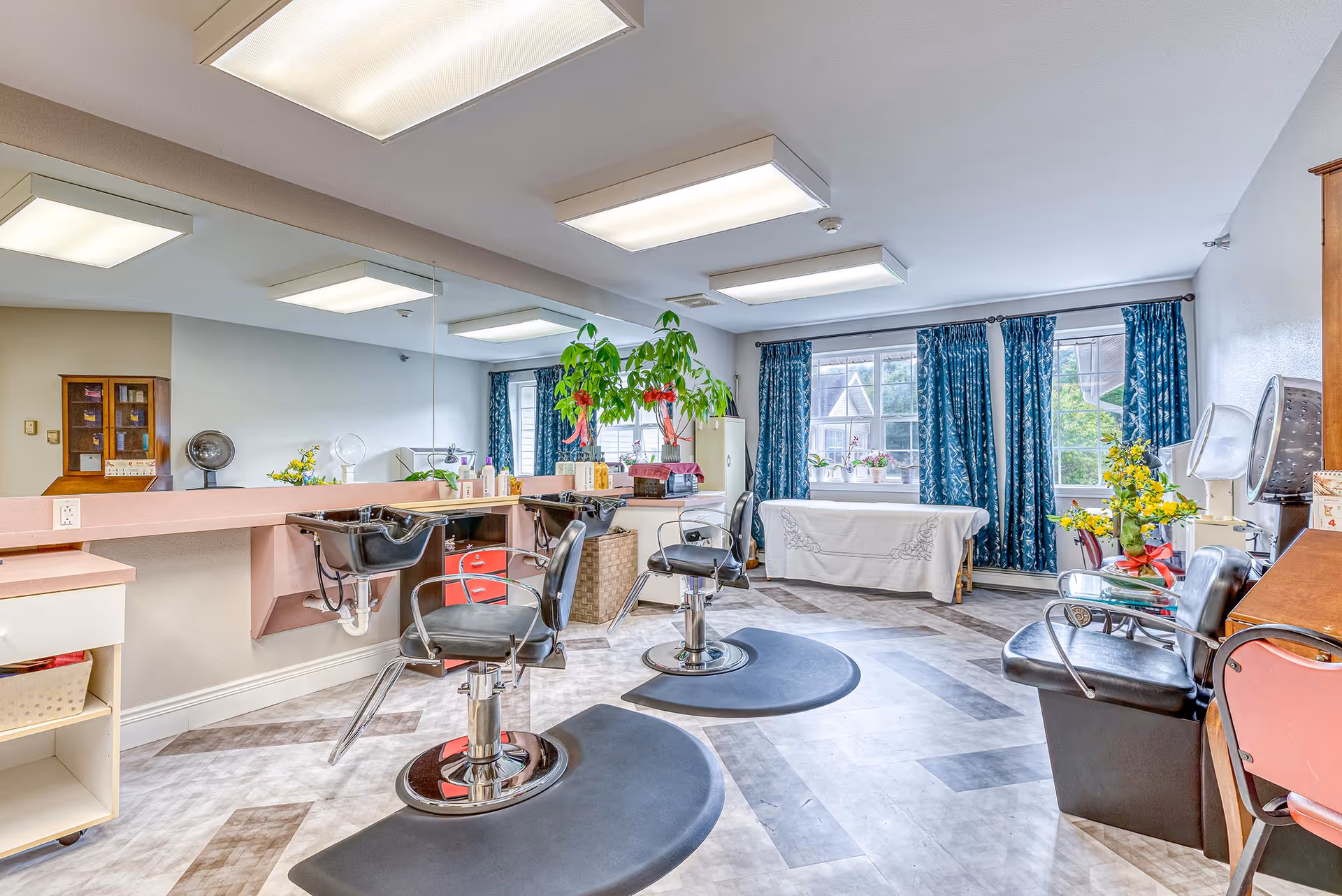 A bright and spacious salon area in a senior living facility with two black salon chairs in front of sinks and a large mirror on the wall. The room has patterned flooring, large windows with blue curtains, plants, and various salon equipment including hair dryers and a table covered with a white cloth.