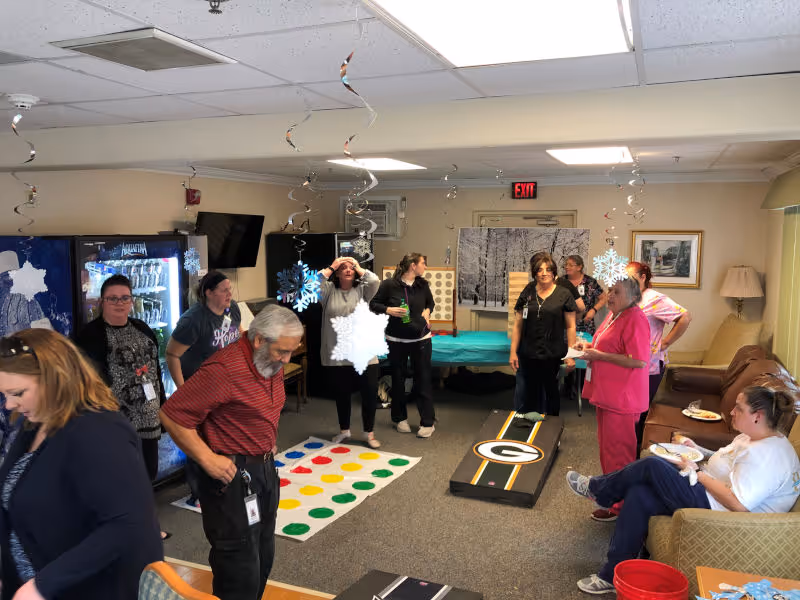 A group of people gathered in a common room decorated with hanging snowflake ornaments. Some individuals are standing and chatting near a Twister game mat and a Green Bay Packers cornhole board, while others are seated on couches eating. The room has vending machines, a television, and framed pictures on the walls.