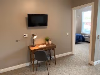 A small workspace with a wooden desk, a modern desk lamp, a small potted plant, and a chair against a beige wall. A flat-screen TV is mounted on the wall above the desk. To the right, there is an open doorway leading to a bedroom with a bed and window visible.