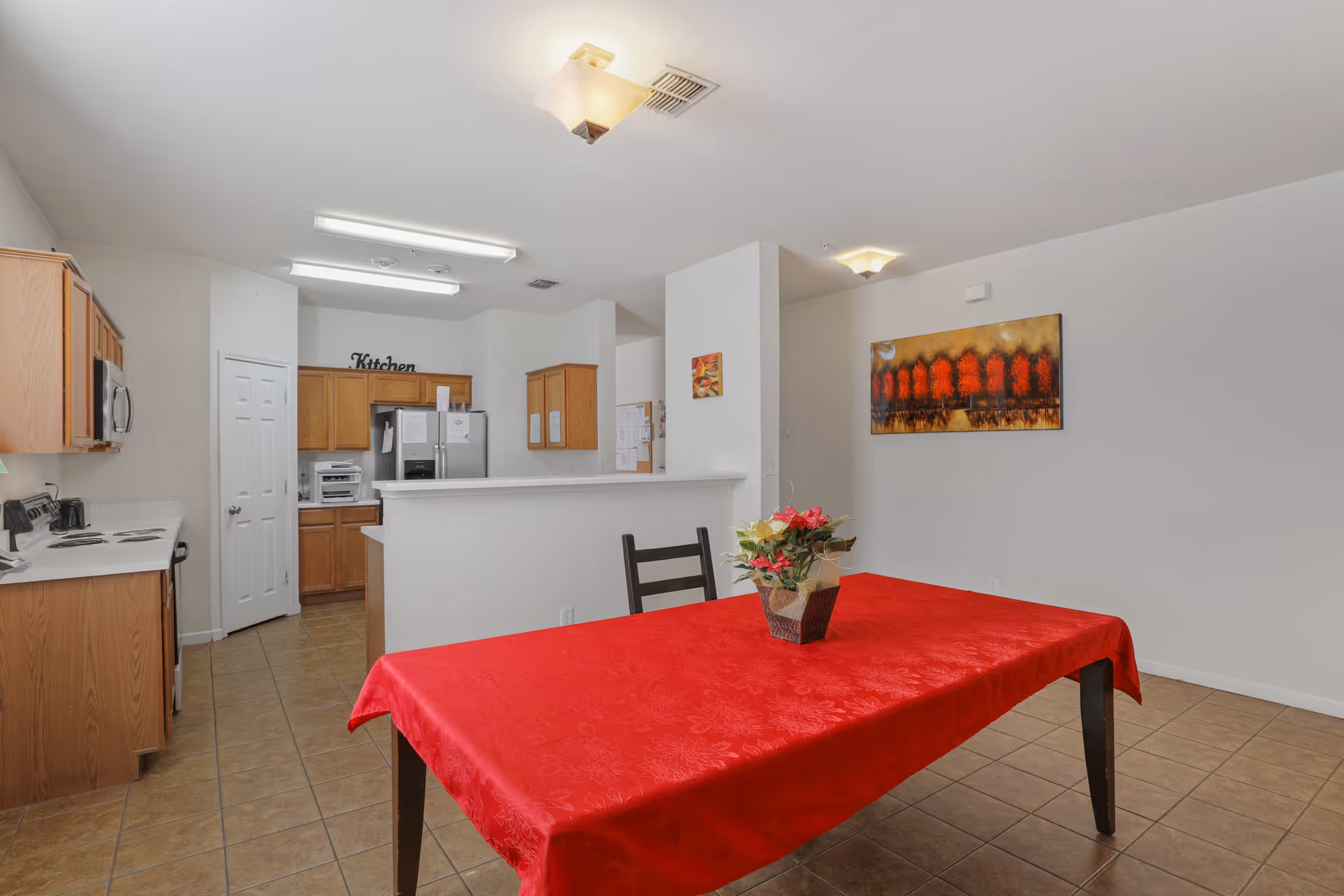 Interior view of a senior living facility kitchen and dining area. The kitchen features wooden cabinets, a refrigerator, microwave, stove, and a white countertop. The dining area has a table covered with a red tablecloth and a small flower arrangement in the center. The walls are white with a colorful painting hanging on one side. The floor is tiled.