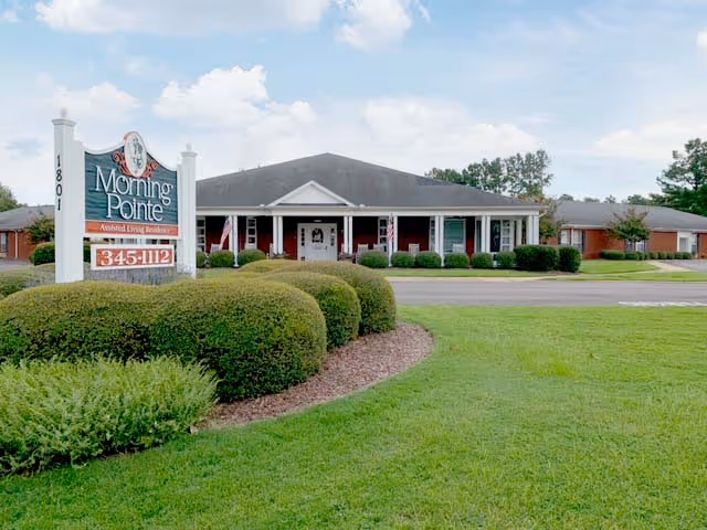 Exterior view of Morning Pointe of Tuscaloosa assisted living residence showing a single-story building with a dark roof, white columns, and a porch. In front of the building is a large sign with the facility name, phone number, and address, surrounded by neatly trimmed bushes and green grass under a partly cloudy sky.
