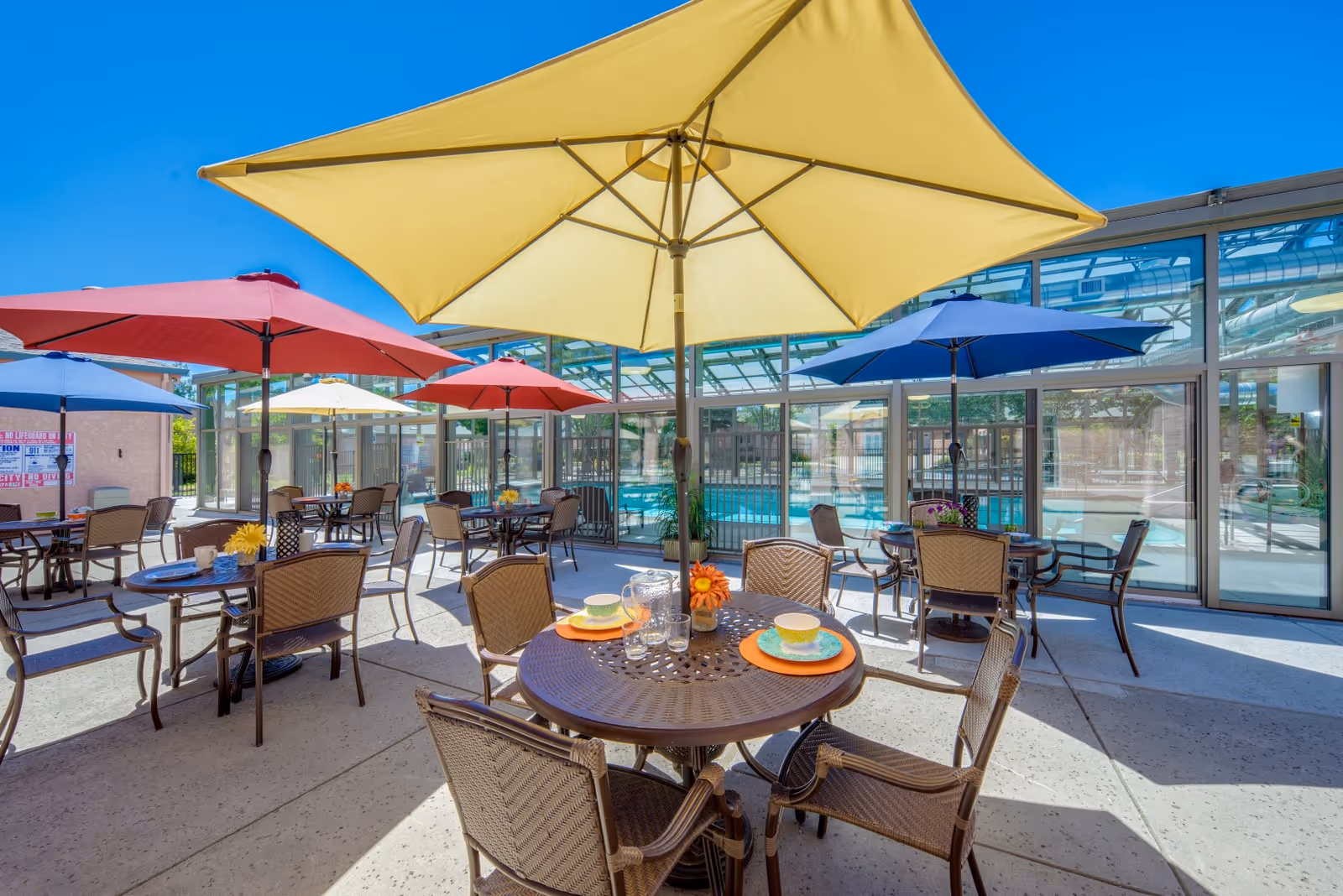 Outdoor patio area with multiple round tables and chairs under large colorful umbrellas including yellow, red, and blue. The patio is adjacent to a glass-enclosed swimming pool area under a clear blue sky.