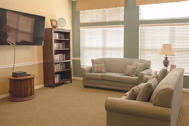 A cozy living room with two beige sofas facing each other, a wooden bookshelf filled with DVDs, a round wooden side table with a cable box, a wall-mounted flat-screen TV, and large windows with blinds letting in natural light. There is also a table lamp on a side table next to one of the sofas.