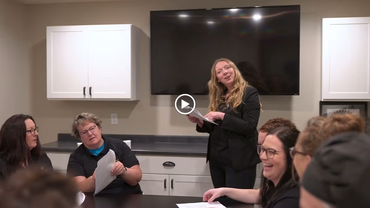 A group of people gathered in a meeting room with white cabinets and a large flat-screen TV mounted on the wall. One woman is standing and speaking while holding papers, and others are seated around a table, some holding papers and smiling.