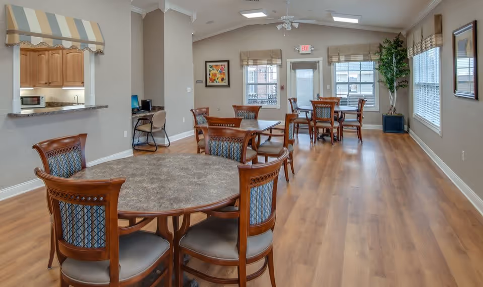 A bright and spacious senior living dining area with multiple round wooden tables and cushioned chairs. The room has large windows with beige blinds, light wood flooring, and neutral-colored walls. There is a small kitchen pass-through window with a striped valance, a computer desk with a chair, and a potted plant in the corner. An exit door is visible at the far end of the room.