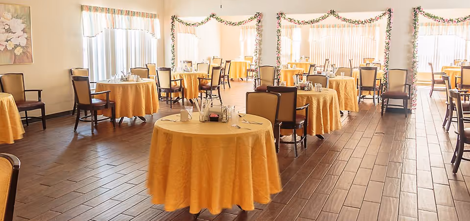 A bright dining room with multiple round tables covered in yellow tablecloths, each set with utensils, napkins, and condiments. The room has wooden flooring, large windows with vertical blinds, and floral garlands decorating the columns. Several wooden chairs with cushioned seats are arranged around the tables.