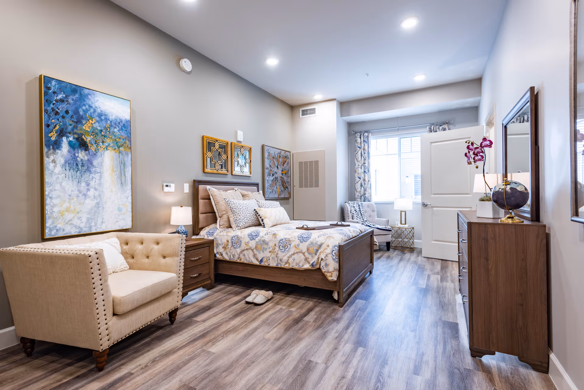 A well-lit senior living bedroom featuring a wooden bed with patterned bedding and multiple pillows, a beige armchair with a pillow, a wooden nightstand with a lamp, two decorative wall mirrors, and artwork on the walls. The room has wood flooring, a large window with curtains, a wooden dresser with a mirror and a purple orchid plant, and a door partially open to another room.