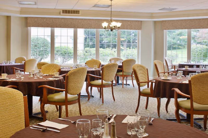 A bright dining room with multiple round tables covered with brown tablecloths, each set with glasses, cups, silverware, and salt and pepper shakers. The room has large windows letting in natural light and offering a view of greenery outside. Comfortable wooden chairs with cushioned seats and backs are arranged around the tables.