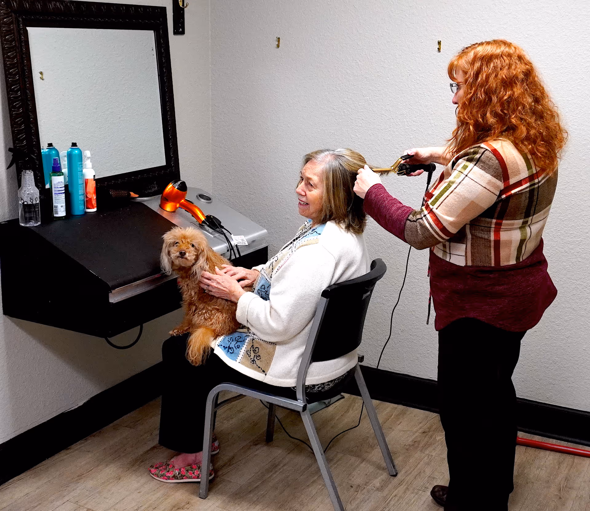 An elderly woman sitting on a chair holding a small brown dog on her lap while another woman with red hair styles her hair using a curling iron in a small room with a mirror and hair care products on a shelf.
