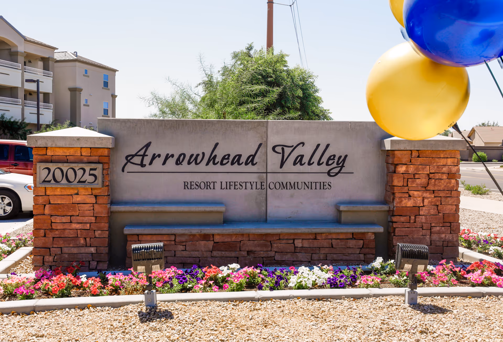 Outdoor stone sign for Arrowhead Valley Resort Lifestyle Communities with the address number 20025 on the left pillar, surrounded by colorful flowers and balloons on the right side. A building and parked cars are visible in the background.