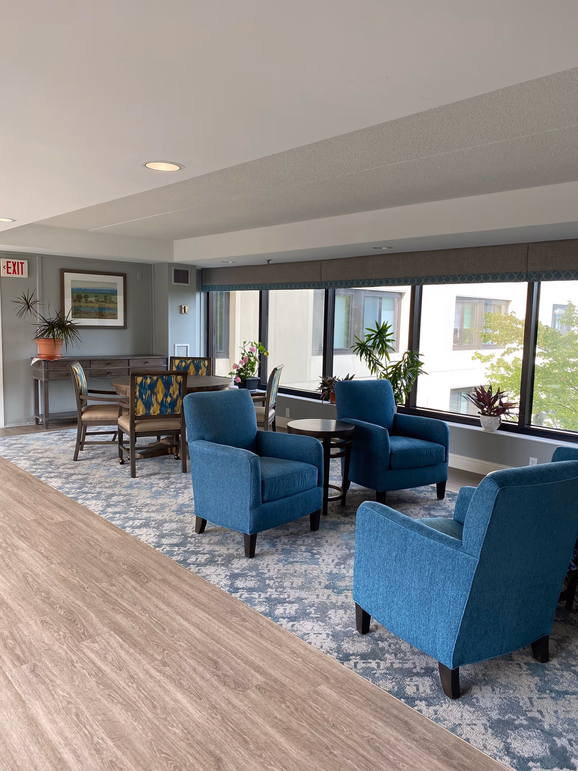 A bright and cozy common area with three blue upholstered armchairs arranged around a small round table. Behind them is a dining table with four chairs, a potted plant on a side table, and large windows letting in natural light. The floor has a combination of wood and patterned carpet, and there is an exit sign on the wall.